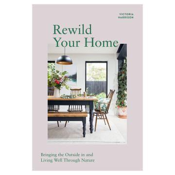 Front cover of Rewild Your Home with a photo of a kitchen table and chairs surrounded by plants, a vase of flowers and a cushion with fern leaf patterns on.