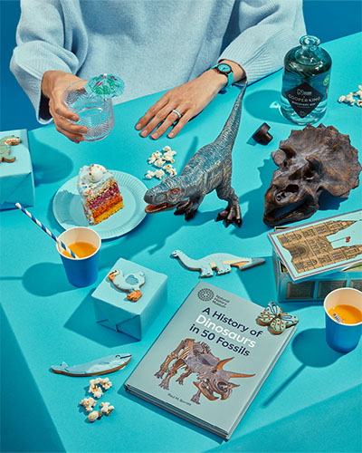 Dinosaur-themed products, including a Triceratops replica skull, a book on fossils and Discovery Gin bottle are arranged on a bright blue table styled as if having a party. A person in a blue jumper sits at the table and only their arms are visible, one is holding a glass.