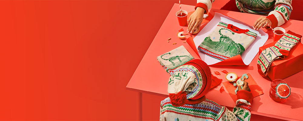 Two people dressed in the Museum Christmas jumper and bobble hat wrap a folded Christmas jumper on a red table, surrounded by red accessories on a red background.
