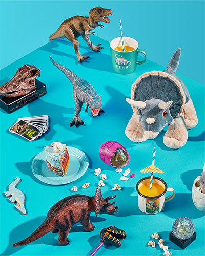 Various dinosaur-themed toys in a party setting against a bright blue background. The toys include T. rex models, a Diplodocus replica skull and a cuddly toy Triceratops.