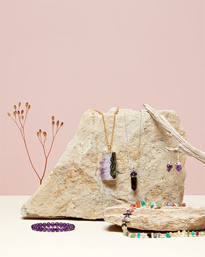 A styled photo of jewellery displayed against a pale pink background. Two necklaces with purple pendants are draped on a piece of rock, next to a pair of earrings hanging on a branch. Below lies a necklace with multi-coloured stone beads and a purple bead bracelet.