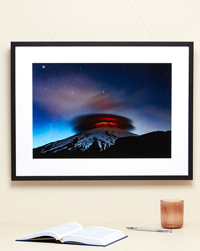 A framed colour photograph of a snow-capped volcano against a dark sky sits on a cream background above an open book and brown cup.