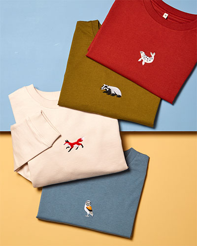 An overhead shot of 4 tops folded and overlapping, laid on a yellow and blue surface. Each top has a motif of an animal on its chest. The red t-shirt’s motif is a seal, the olive-green t-shirt’s is a badger, the cream jumper’s is a fox and the blue t-shirt’s is an owl.